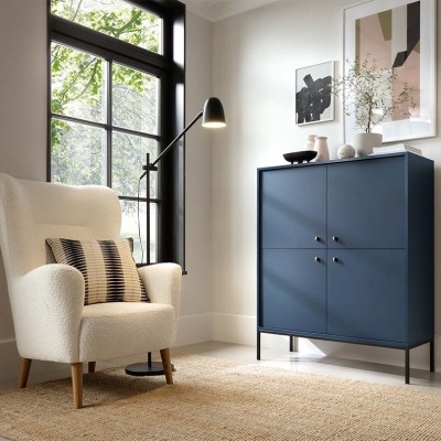 Poltrona branca, candeeiro de pé, armário azul escuro com objetos decorativos e pinturas abstratas numa sala clara