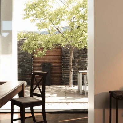 Sala de estar com mesa e cadeiras de madeira escuras junto a porta de vidro aberta para área externa com árvore e muro de pedra