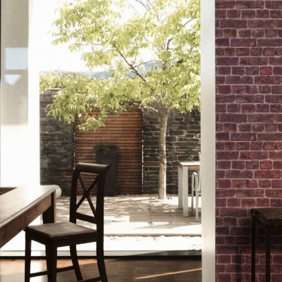Sala com mesa e cadeira de madeira e parede de tijolos vermelhos, aberta para área externa com árvores.