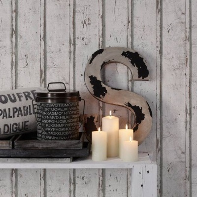 Velas acesas, jarra preta com texto, almofada branca com texto e letra decorativa 'S' numa mesa contra parede de madeira branca