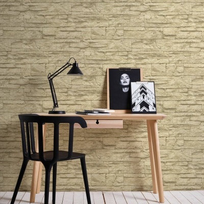 Mesa de escritório de madeira clara com cadeira preta, lâmpada de mesa, quadros decorativos e parede de pedra bege.