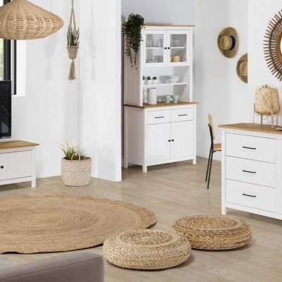 Sala de estar com móveis brancos e tampo de madeira, espelho redondo, tapete e pufes de ráfia, candeeiro de vime