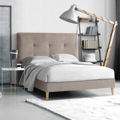 Cama com cabeceira estofada bege, roupa de cama branca, mesa de apoio com candeeiro, estante com livros