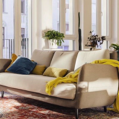 sofá bege com almofadas e manta amarela numa sala iluminada com plantas e estante