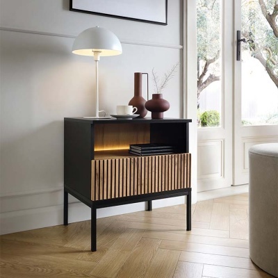 Mesa de cabeceira preta com porta de madeira aos ripados e objetos decorativos numa sala com porta de vidro.