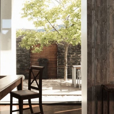 Mesa e cadeira de madeira em sala rústica com parede de tábuas escuras e área exterior com árvore e parede de pedra