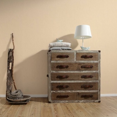 Cómoda de madeira com seis gavetas, almofadas cinzentas e candeeiro branco num ambiente de madeira clara e parede bege