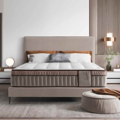 Cama de casal estofada cinza claro com colchão branco, almofadas castanhas e cinza, candeeiros brancos, mesinhas brancas e pufe cinza com tecido castanho