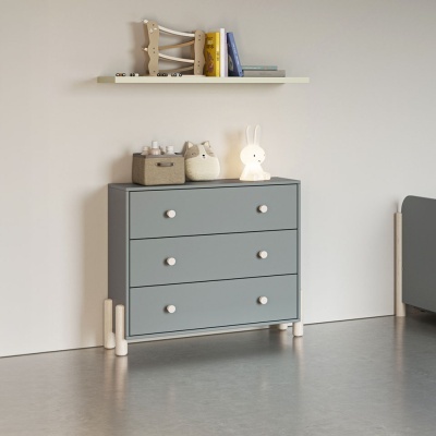 Cómoda cinza com três gavetas e puxadores brancos, prateleira branca com livros e brinquedos, luz decorativa de coelho.