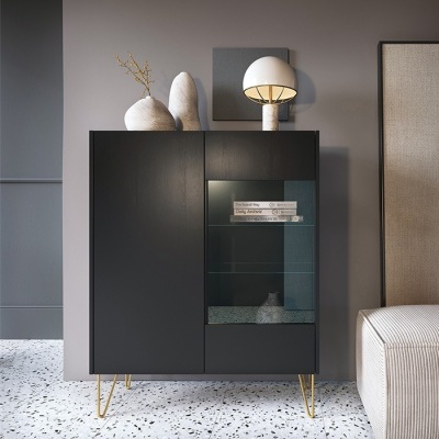 Armário preto com porta de vidro e pernas douradas, decorado com vaso e candeeiro, em ambiente interior moderno.