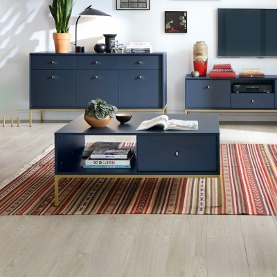 Sala com móveis azuis escuros e pés dourados, mesa de centro, aparador, estante, plantas e tapete listrado colorido