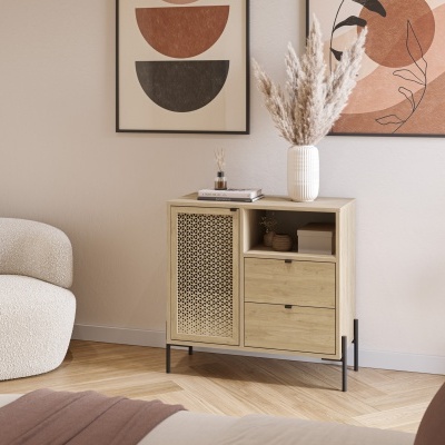 Cómoda de madeira clara com porta com padrão geométrico e três gavetas, vaso branco com flores secas em cima, quadros abstratos na parede