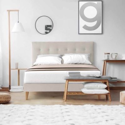 Quarto com cama estofada bege, abajur de madeira, relógio, quadro letra g, mesa e banco de madeira com almofadas e tapete branco