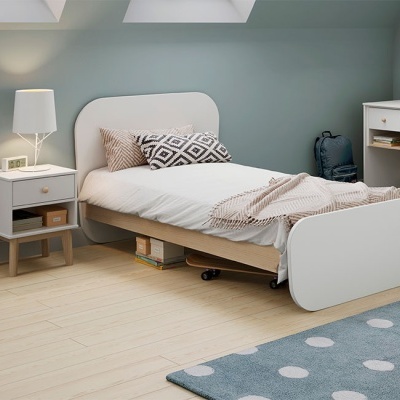 Cama solteiro branca com almofadas decorativas e manta bege num quarto com mesa de cabeceira e tapete azul com bolinhas