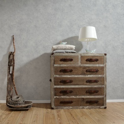 Cómoda rústica de madeira com seis gavetas, livros e abajur branco junto a parede cinzenta