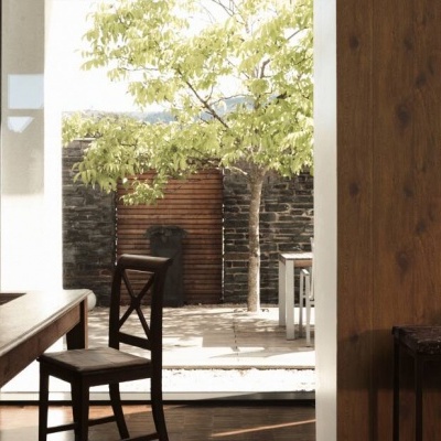 Mesa e cadeira de madeira escura numa sala com porta aberta para pátio com árvore e muro de pedra