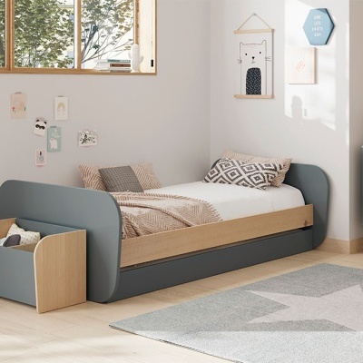 Cama infantil cinzenta com roupa de cama bege e almofadas geométricas num quarto branco com tapete cinzento estrelado.