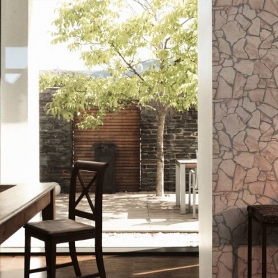 Sala de jantar com parede de pedra clara, mesa e cadeira de madeira, consola preta, porta aberta para pátio com muro de pedra escura e árvore