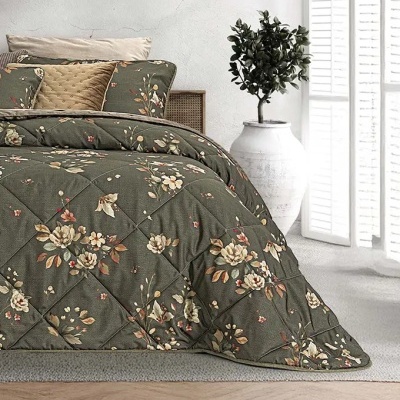 Edredão floral verde escuro numa cama com almofadas castanhas e bege, junto a vaso branco com planta e janela persiana.