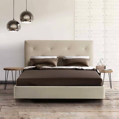 Cama de casal estofada bege com roupa castanha, mesas de madeira e candeeiros suspensos metálicos
