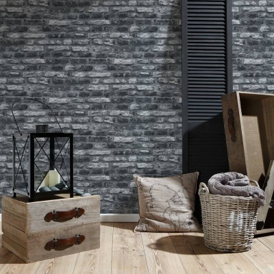 Conjunto decorativo com caixa de madeira, lanterna preta, almofada, cesta de vime com manta, e suporte de madeira junto a parede de tijolos cinzentos.