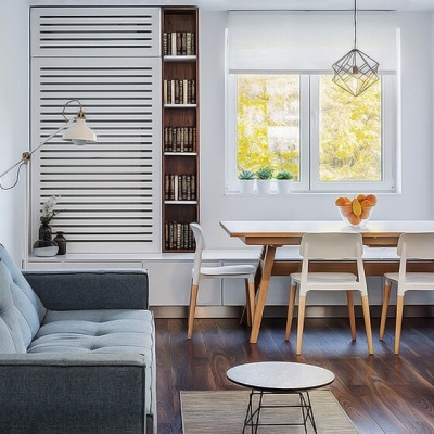 Sala de estar com sofá cinza, mesa de jantar em madeira clara, cadeiras brancas e estante com livros
