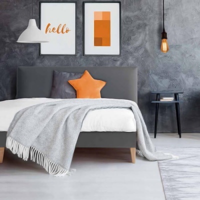 Quarto com cama cinza, manta cinza clara, almofada laranja estrela, candeeiros e quadros na parede.