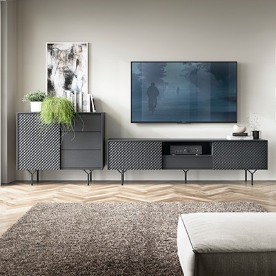 Móvel cinzento texturizado com padrão geométrico e três gavetas alinhado a parede clara com decoração de plantas e quadro.