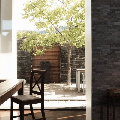 Sala com parede de tijolos cinza escuro, mesa e cadeira de madeira, com vista para pátio com árvore