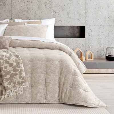 Cama com colcha bege texturizada, almofadas brancas, manta com padrão de folhas, parede de pedra cinza e móvel cinza