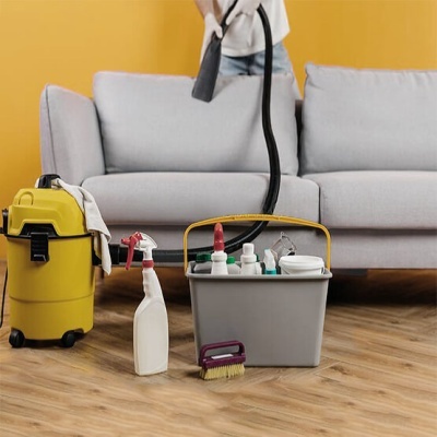 Sofá cinzento claro com aspirador preto e amarelo, balde cinza com produtos de limpeza e pincel no chão de madeira