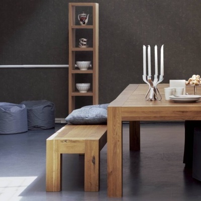 Sala de jantar com mesa e banco de madeira clara, candelabro prateado e estante alta com pratos e vasos brancos numa parede escura