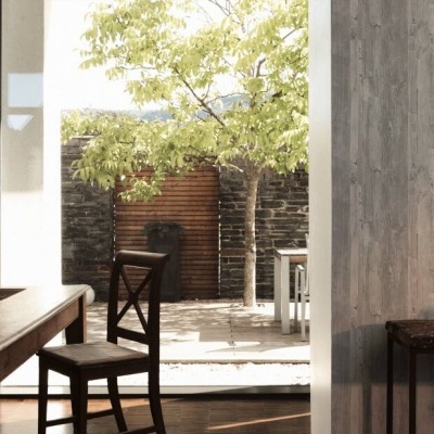 Sala de jantar com mesa e cadeiras em madeira escura junto a uma janela grande aberta para um pátio com árvore