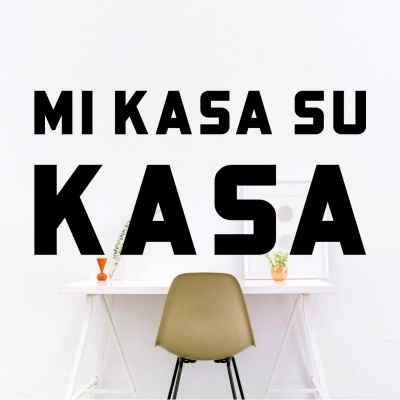 Texto em preto 'MI KASA SU KASA' numa parede branca acima de uma secretária branca com cadeira verde oliva.