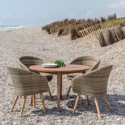 Conjunto de mesa redonda em madeira com quatro cadeiras de vime sintético bege, numa praia de seixos com cerca de madeira ao fundo