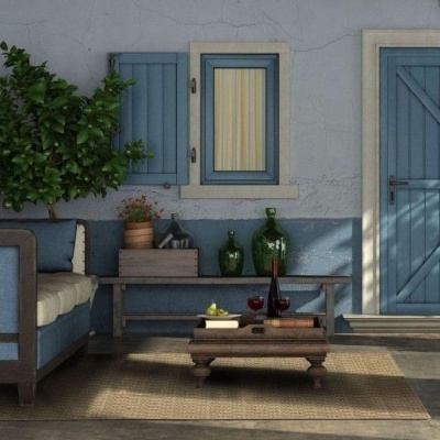 zona de estar exterior com sofá azul e almofadas brancas, mesa de centro em madeira com vinho e fruta, jarras verdes, planta, janela e porta azuis