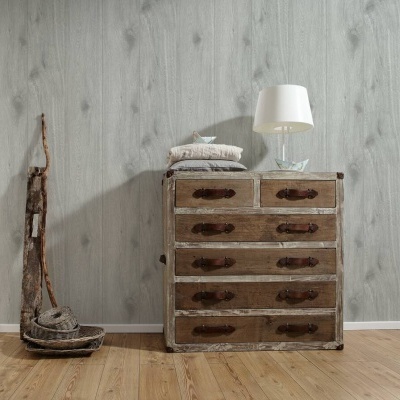 Cómoda de madeira envelhecida com cinco gavetas e candeeiro branco sobre ela, piso de madeira clara e parede cinzenta com padrão rústico.