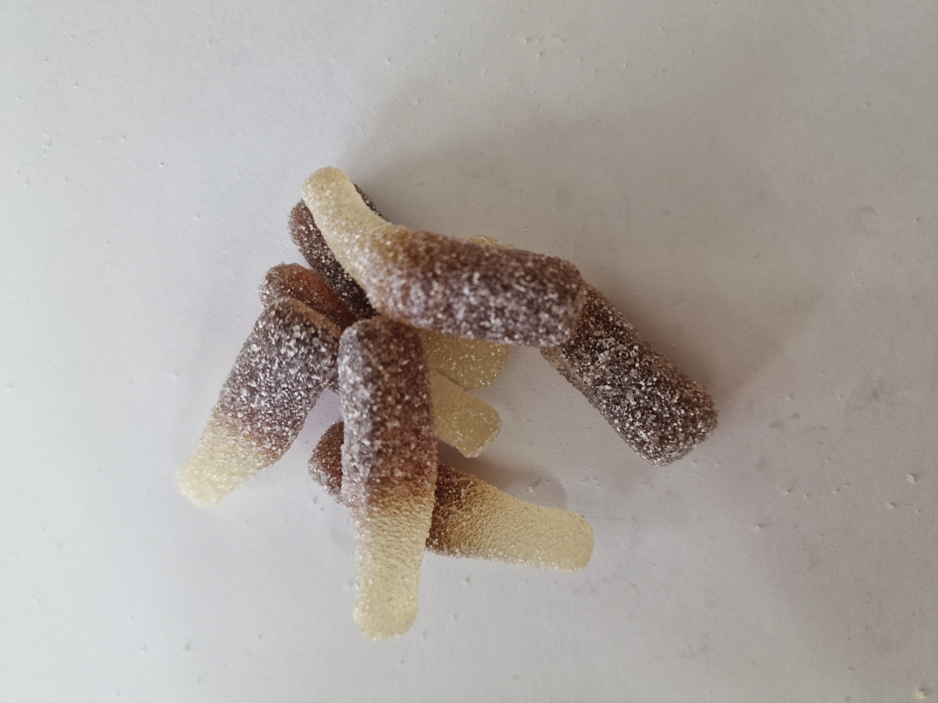 Gomas em forma de garrafa castanhas e amarelas com açúcar, sobre superfície branca.