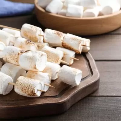 Espetadas de marshmallows tostados em tábua de madeira com tigela de marshmallows brancos ao fundo
