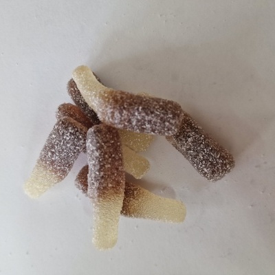 Gomas em forma de garrafa castanhas e amarelas com açúcar, sobre superfície branca.