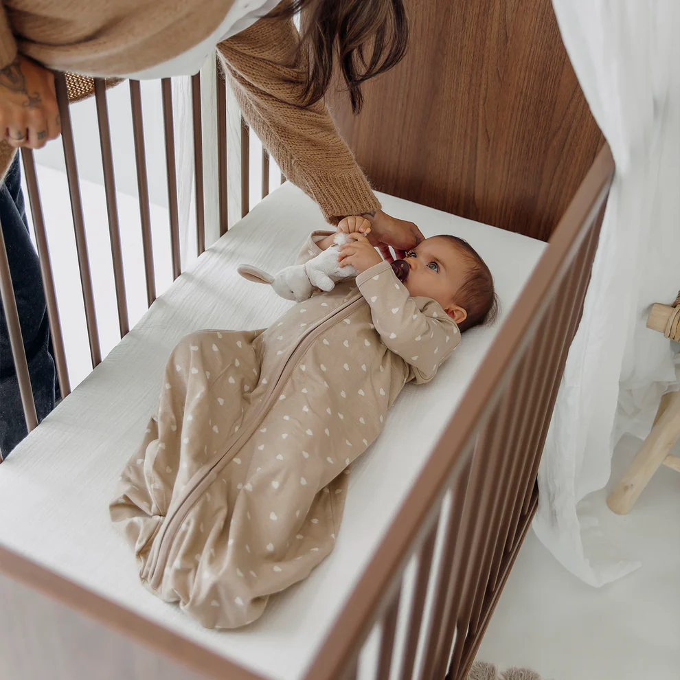 Saco de dormir com Mangas TOG 1.0 Sweet Heart Sand | Meyco Baby Bebé em saco de dormir bege com corações brancos em mini berço de madeira castanha