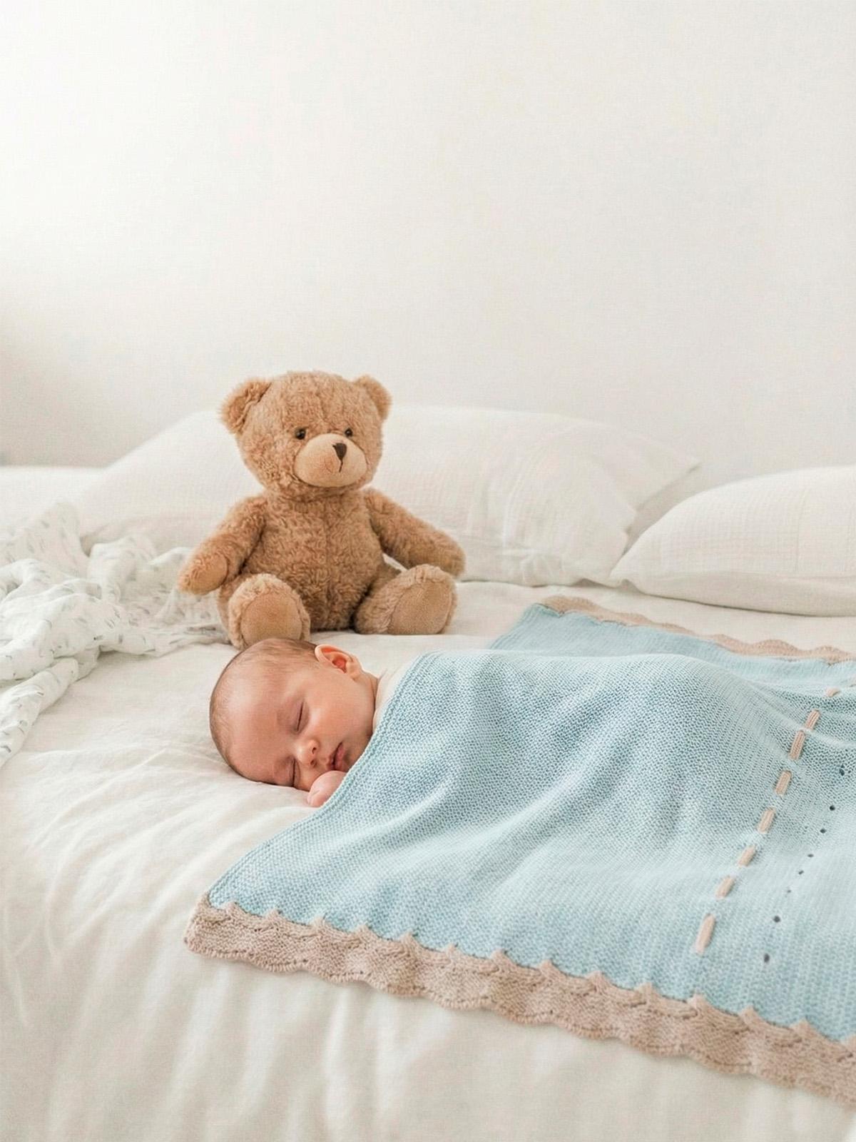 Manta malha na cor Iconic da Mac Ilusión - Coleção Abetos Iconic Bebé adormecido coberto por cobertor azul com urso de peluche castanho na cama branca.