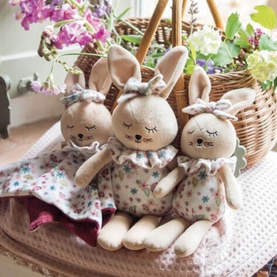 Três coelhinhos de pelúcia com vestidos floridos sentados junto a uma cesta de flores