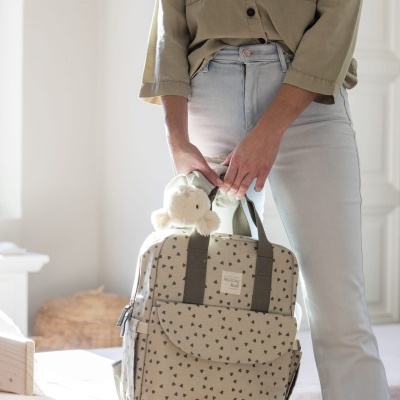 Mochila bege com padrão de triângulos pretos e peluche branco, segurada por pessoa com camisa bege e calças claras num quarto