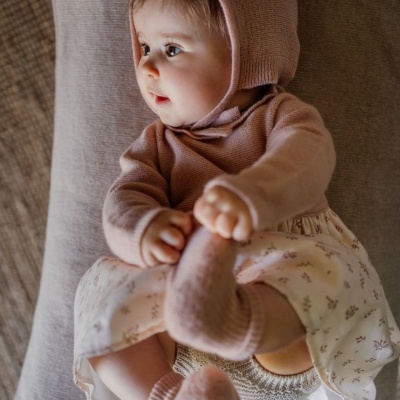 Bebé vestido com macacão e touca rosa, vestido branco com flores, meias rosa, sentado numa cadeira cinzenta