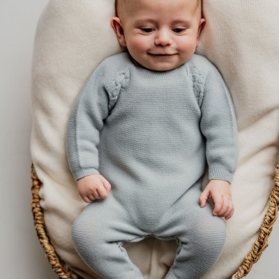 Bebé em macacão azul claro de malha, deitado numa almofada creme dentro de um cesto de verga