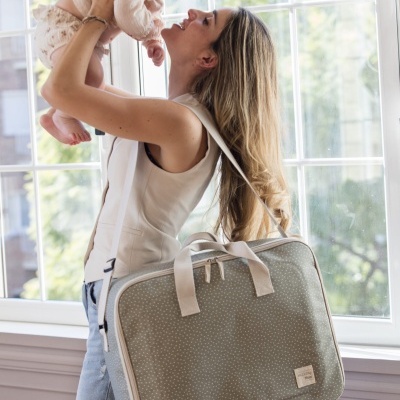 Mulher segurando bebé perto de janela com mala verde-acinzentada e pontos brancos