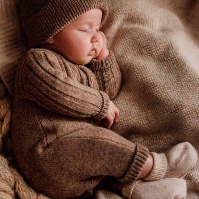 Bebé a dormir com pijama e gorro de malha castanhos, meias brancas e manta bege tricotada