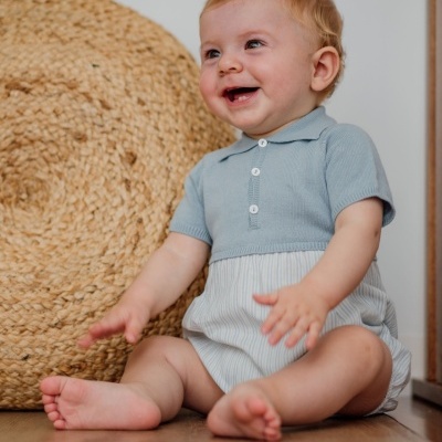 Bebé com roupa azul e branca sentado junto a peça de ráfia redonda no chão de madeira