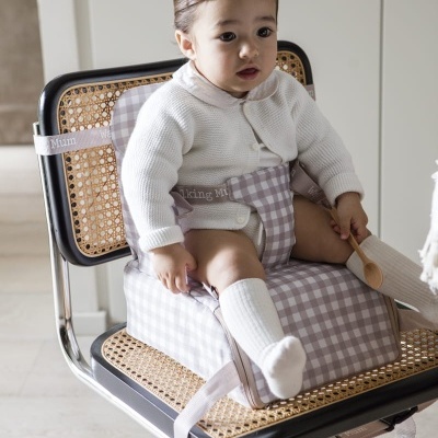 Bebé sentado em almofada de elevação cinza com padrão de quadrados numa cadeira com estrutura metálica e assento em vime.